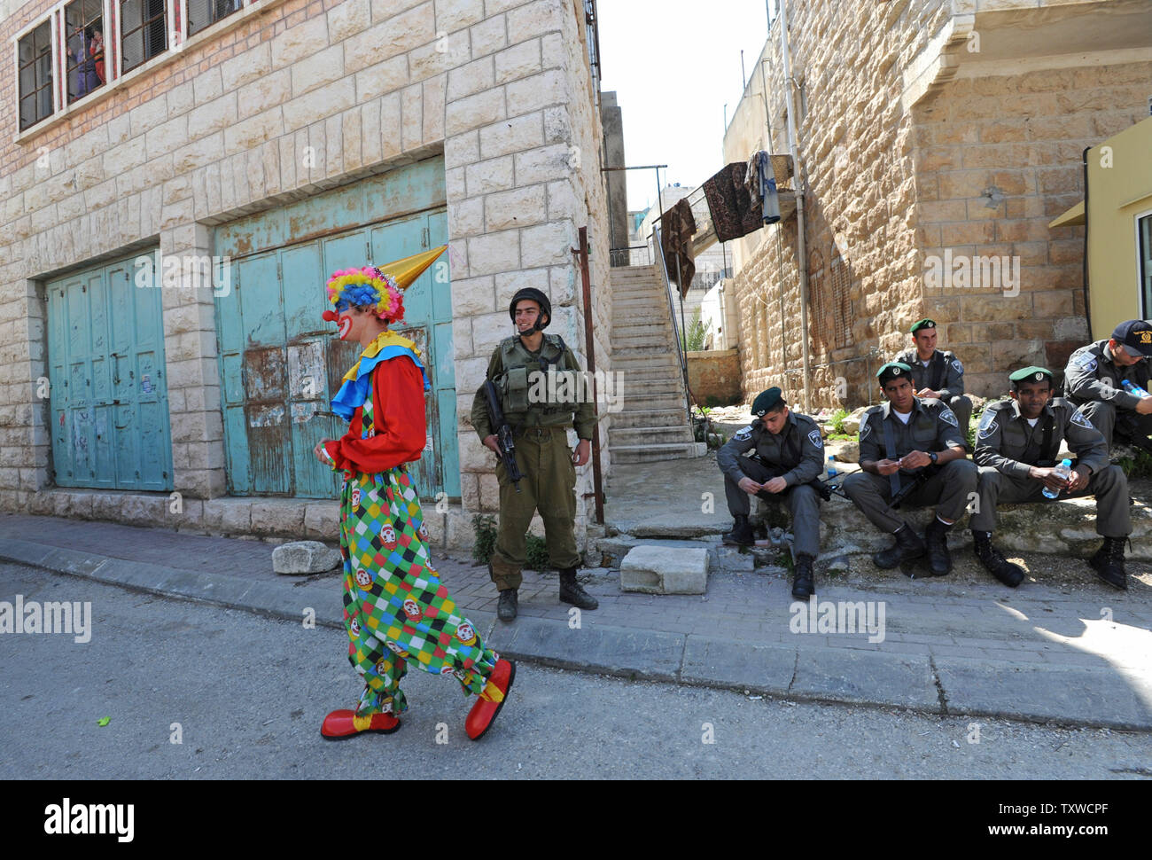 A Jewish settler, dressed in a clown costume, walks by Israeli soldiers waiting to secure the area before  the annual parade marking the Jewish holiday of Purim, in the divided West Bank town of Hebron, Thursday, March 8, 2012. The festival of Purim commemorates the rescue of Jews from genocide in ancient Persia as told in the book of Esther.  UPI/Debbie Hill Stock Photo