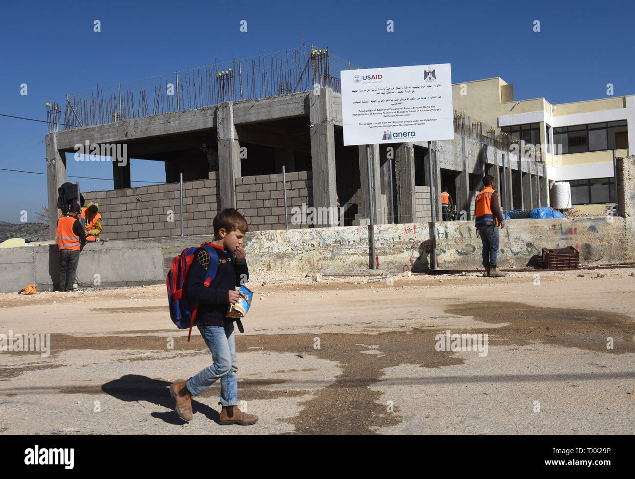 A Palestinian boy eats while walking by construction workers renovating and building an extension to the girl's school in Al-Jabba, near Bethlehem, West Bank, which is funded by USAid, United States Agency for International Development, January 23, 2019. The $1.4 million dollar project will be terminated at the end of January because U.S. President Donald Trump's administration will stop funding Palestinian projects and also because Palestinian Prime Minister Rami Hamdallah has stated that the Palestinian Authority will cease accepting funding from America. The school will be unusable for the Stock Photo