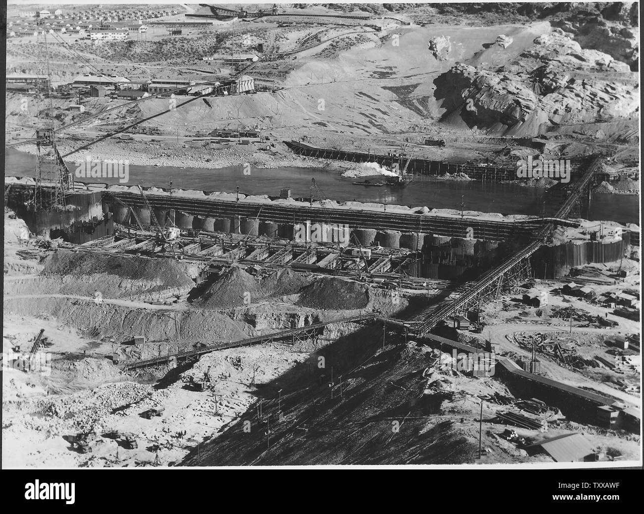 Behind section E of west cofferdam where bedrock overburden is being removed for placing of block 40 of the Grand Coulee dam. Across the river are the contractor's machine shops, above which is the general office building , mess hall, the 64 one-room houses, and the hospital - the east cofferdam and conveyor to the west side spoil pile.; Scope and content:  Photograph from Volume Two of a series of photo albums documenting the construction of the Grand Coulee Dam and related work on the Columbia Basin Project. Stock Photo