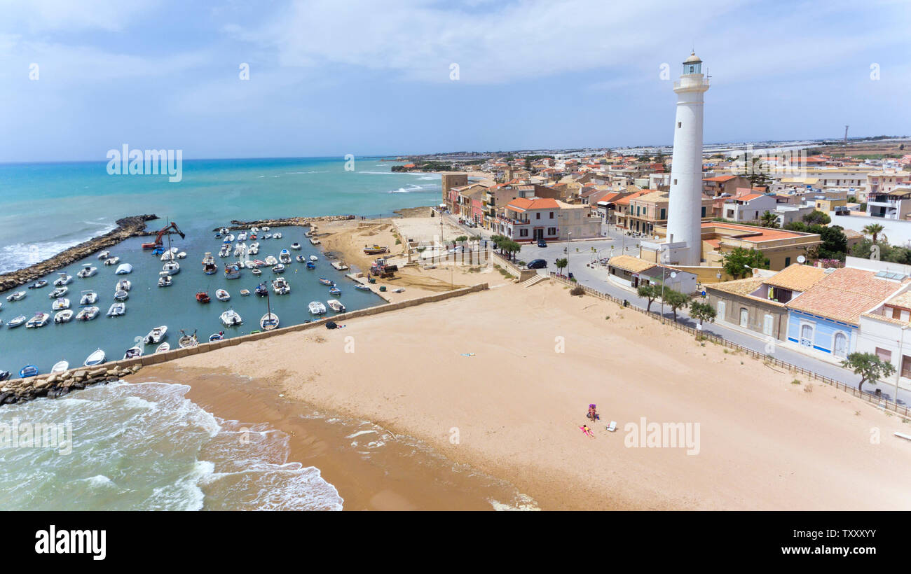 Golden sandy beach with shallow waters, small port with boats, white tall lighthouse in Sicilian fishing village of Punta Secca, set of TV series Insp Stock Photo