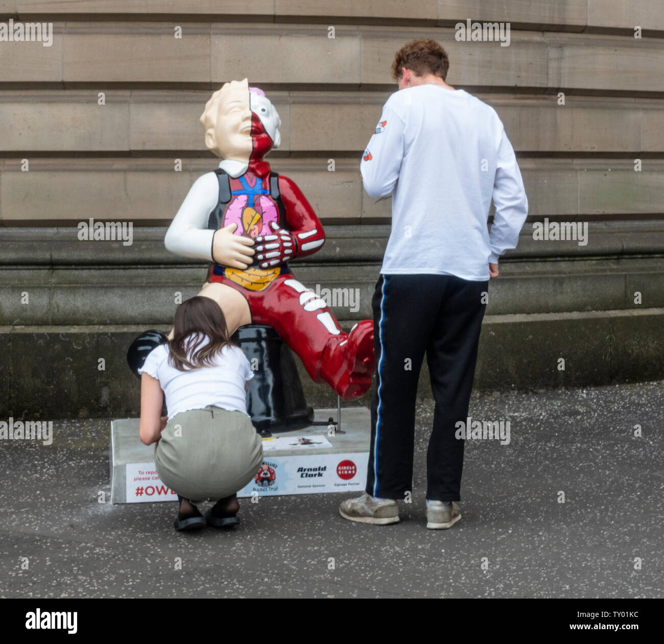 A young couple examine Anatomical Boy by Simon Messer, part of the Oor Wullie Big Bucket Trail 2019. Outside the Mitchell Library, Glasgow, Scotland Stock Photo