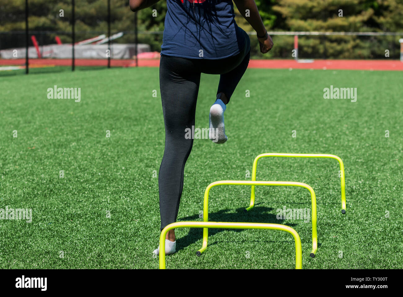 A high school girl is performing speed and agility drills over hurdles with no shoes on, only socks on a green turf field wearing black spandex. Stock Photo
