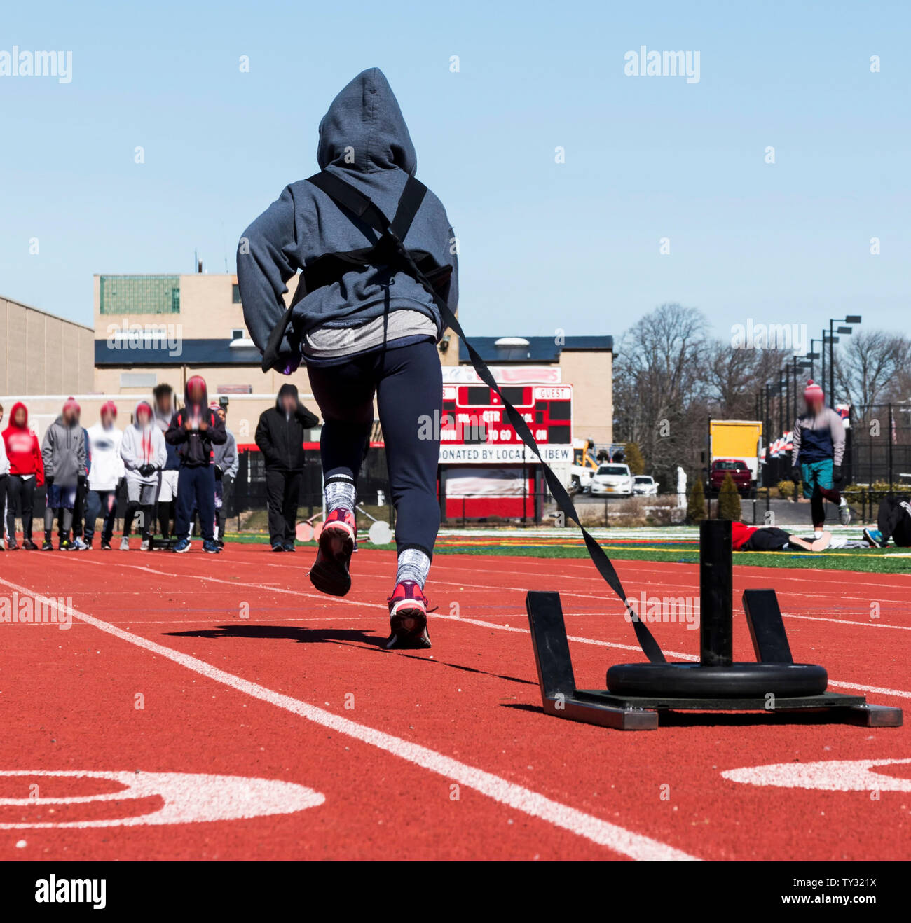 A track and field athlete is pulling a sled with 25 pounds on it down a red track as his teammates watch in the distance. Stock Photo