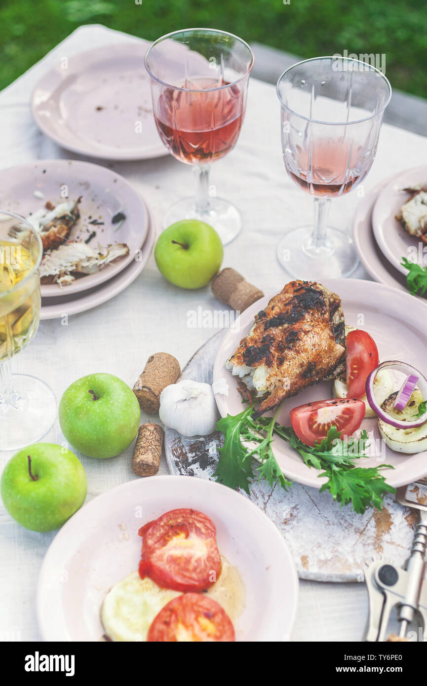 Glasses of white and rose wine, grilled fish plates, vegetables, salad and fruits on the table. Summer party in the backyard. Vertical shot Stock Photo