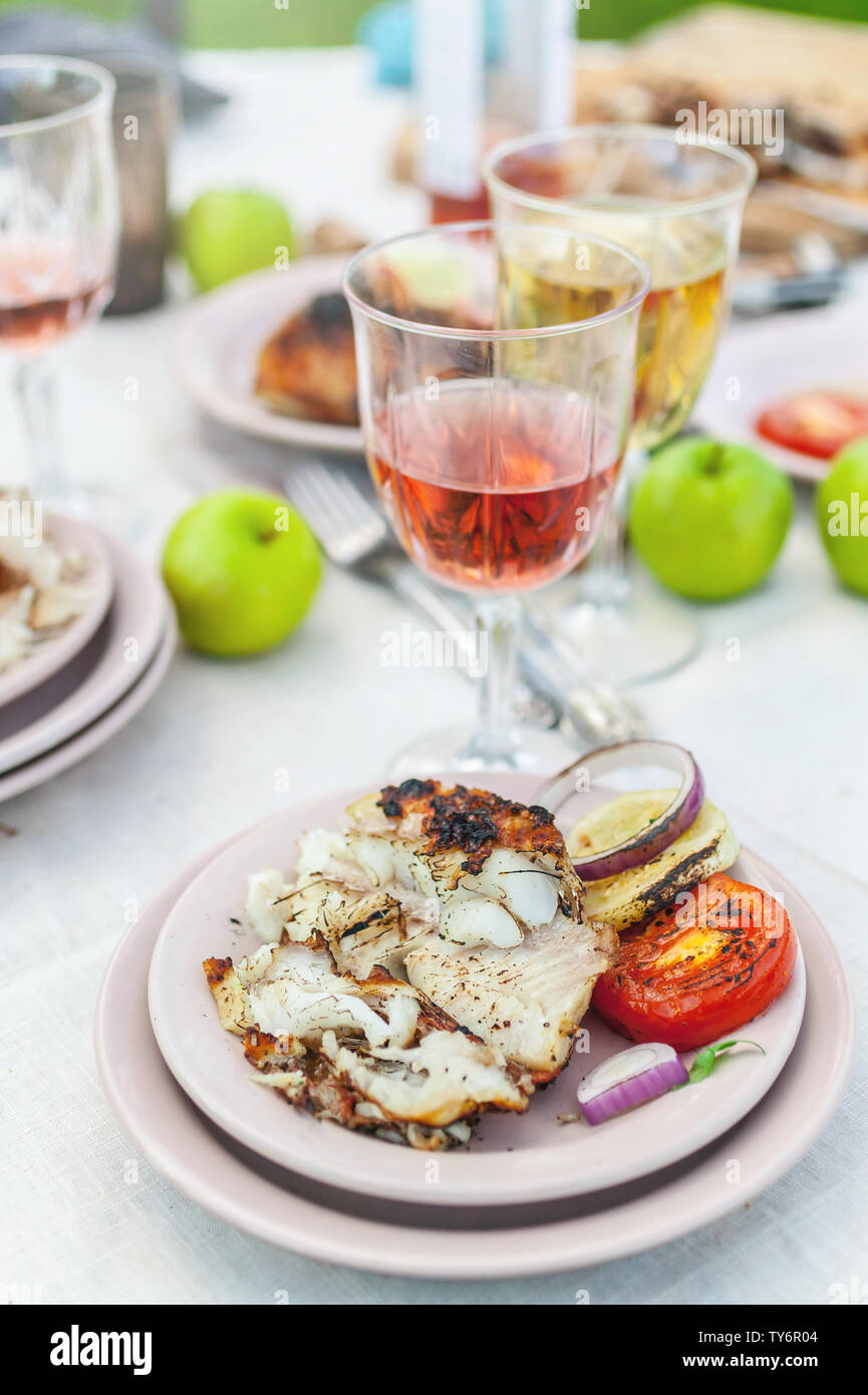 Grilled grouper, glasses with white and rose wine, plates, vegetables, salad and fruits on the table. Summer party in the backyard. Vertical shot Stock Photo