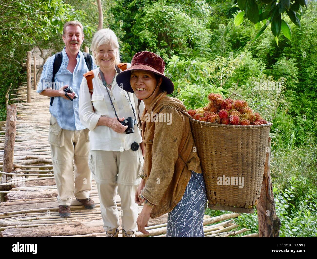 A smiling European couple pose for photo on bridge in countryside with friendly Vietnamese woman carrying basket of rambutan fruit on her back Stock Photo