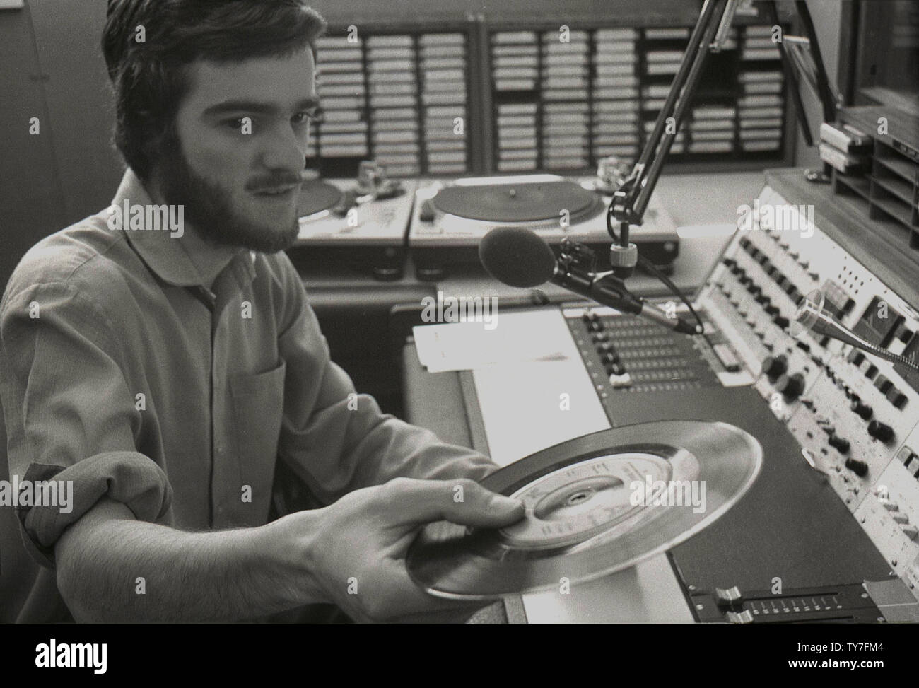 1980s, a young male DJ for a local radio station sitting at his control desk or booth with microphone, holding a gramophone record. The turntables to play the disc can be seen in the picture, England, UK. Stock Photo