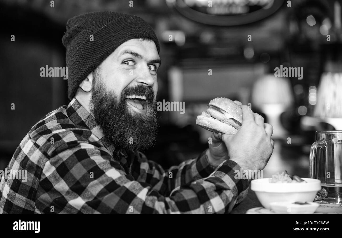 Hipster hungry man eat burger. Man with beard eat burger menu. Brutal hipster bearded man sit at bar counter. High calorie food. Cheat meal. Delicious burger concept. Enjoy taste of fresh burger. Stock Photo