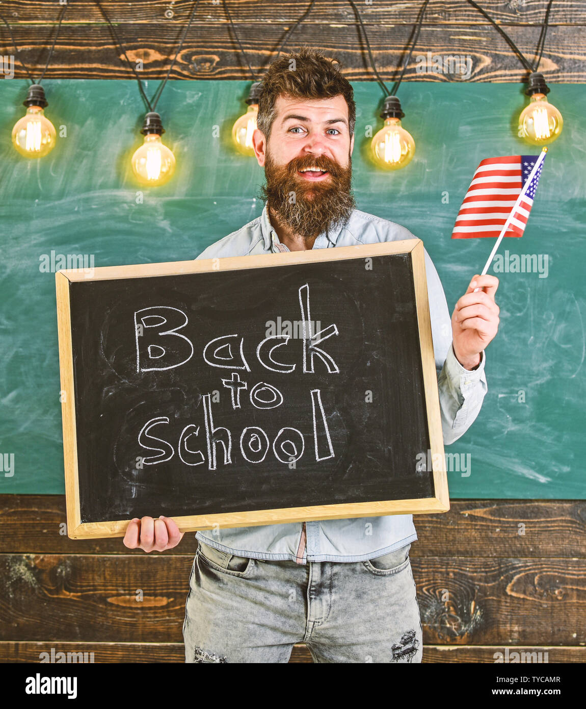 Man with beard on smiling face welcomes to american school, chalkboard on background. American school concept. Teacher holds blackboard with written phrase back to school and flag of USA. Stock Photo