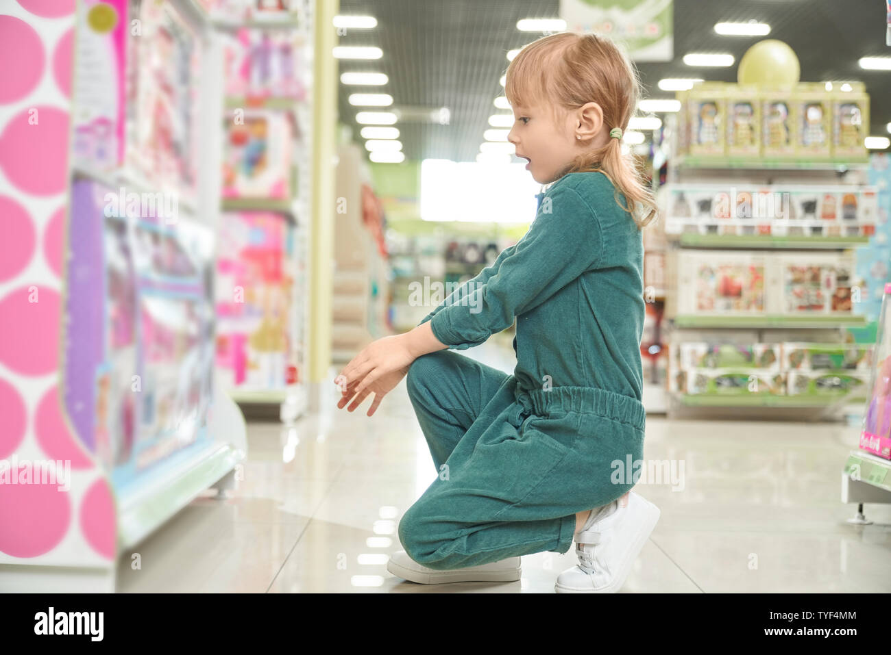 Amazed kid sitting near stands with new toys and choosing beautiful dolls in big shop. Happy girl with open mouth buying presents and gifts in mall. Concept of shopping and playing. Stock Photo