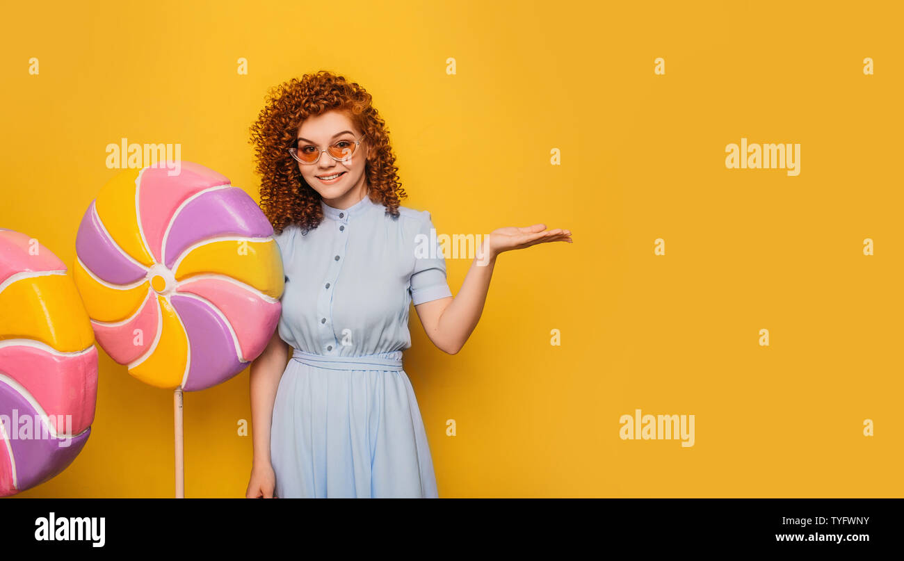 curly red-haired positive woman with lollipops pointing hands into empty yellow background Stock Photo