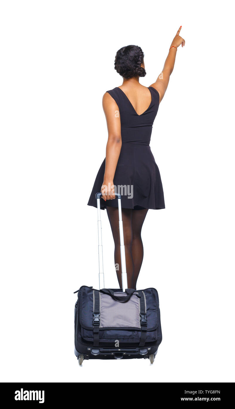 Back view of a black African-American walking with a suitcase and pointing hand forward. Tanned girl in a short dress rolls a suitcase. beautiful girl Stock Photo