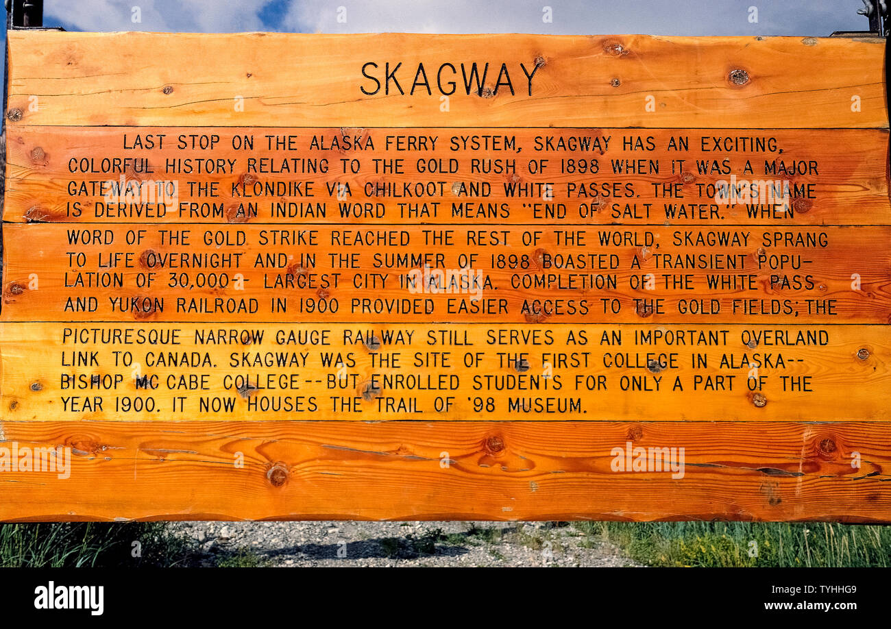 An outdoor wooden sign describes a brief history of Skagway, Alaska, USA, a remote outpost made famous by the 1897-98 gold rush to the Klondike in the Yukon Territory of Canada. Today it is a popular tourist attraction reached by cruise ships and the Alaska ferry system that provides a marine highway to communities in Southeast Alaska that have no access by road. Many visitors to Skagway board the historic gold-rush White Pass & Yukon Railway for a scenic mountainous train excursion that climbs from sea level to the 2,915-foot (888-meter) summit of White Pass. Stock Photo