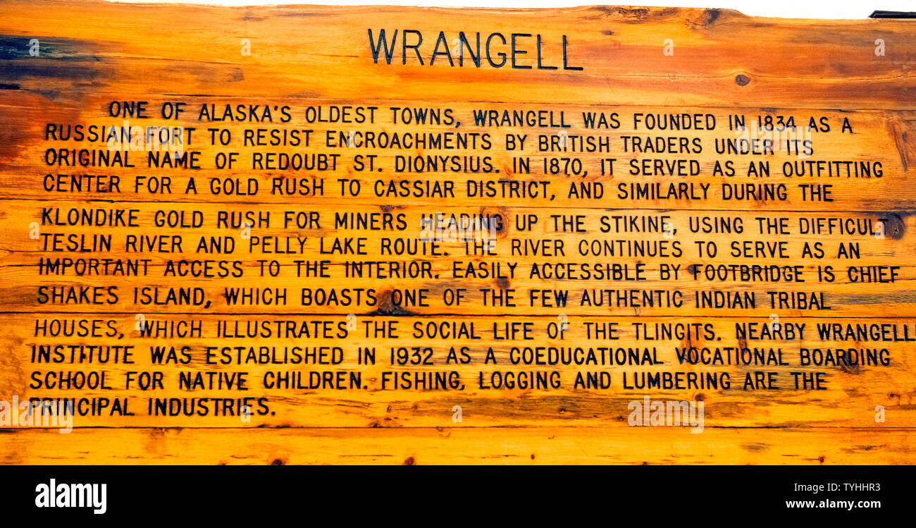 An outdoor weather-worn wooden sign explains the early history of Wrangell, one of the oldest towns in Alaska, USA. It began in 1834 when Fort Dionysius was built by the Russians to protect their interests in the sea otter fur trade. Wrangell later became a supply center for prospectors using the Stikine River to reach goldfields in Canada, including the Klondike. The town also has a longtime history with the Native Tlingit Indians that tourists can share by visiting Shakes Island to see hard-carved totem poles and a recently restored tribal house. Stock Photo
