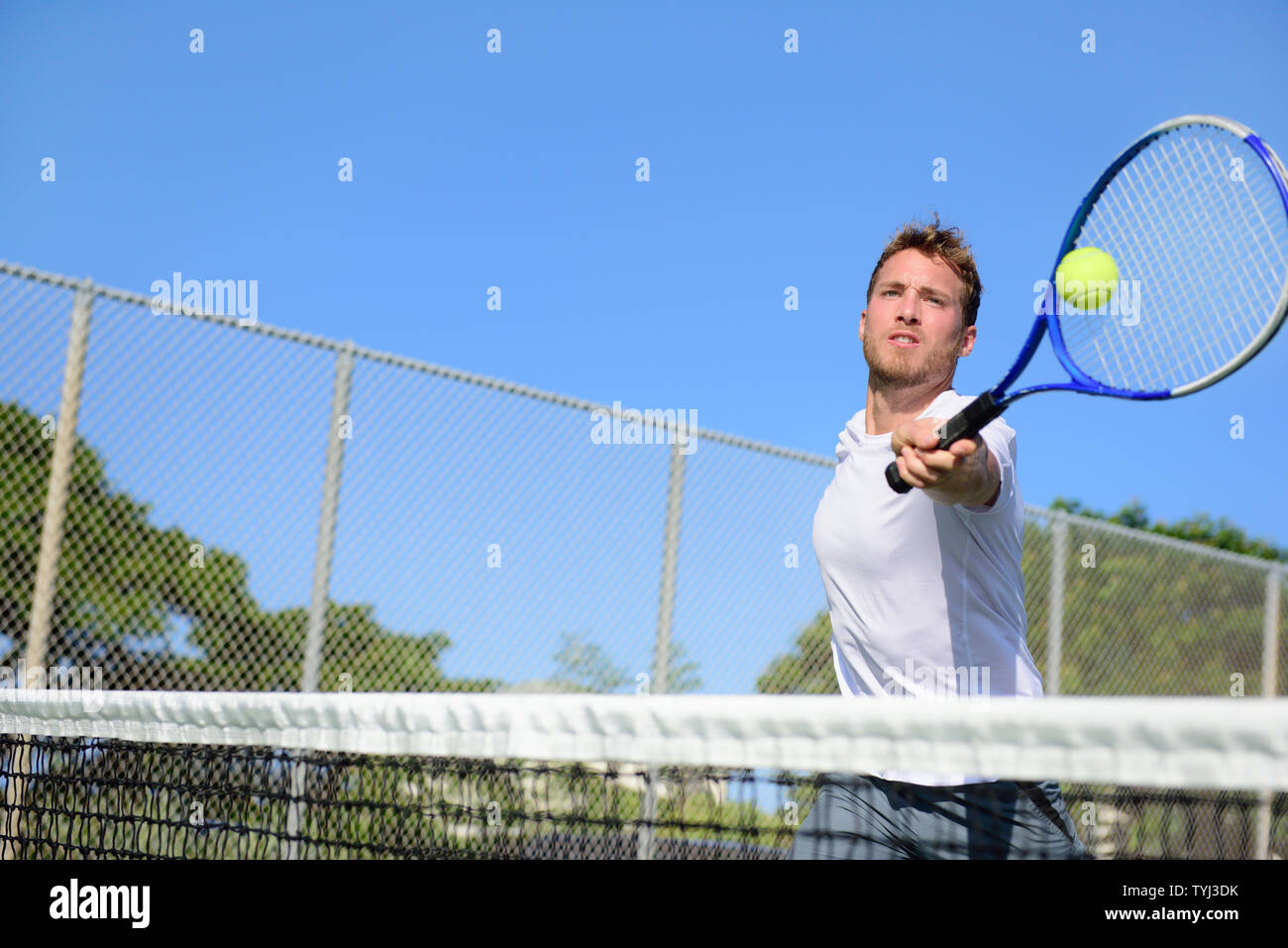 Tennis player man hitting ball in a volley. Male sport fitness athlete playing tennis on outdoors hard court in summer. Healthy active lifestyle concept. Stock Photo