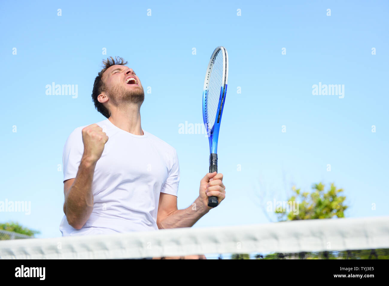 Tennis player man winning cheering celebrating victory. Winner man happy in celebration of success and win. Fit male athlete on tennis court outdoors holding tennis racket in triumph by the net. Stock Photo