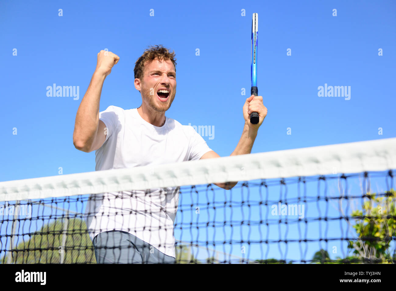 Tennis player celebrating victory. Winning cheering man happy in celebration of success and win. Fit male athlete winner on tennis court outdoors holding tennis racket in triumph by the net. Stock Photo