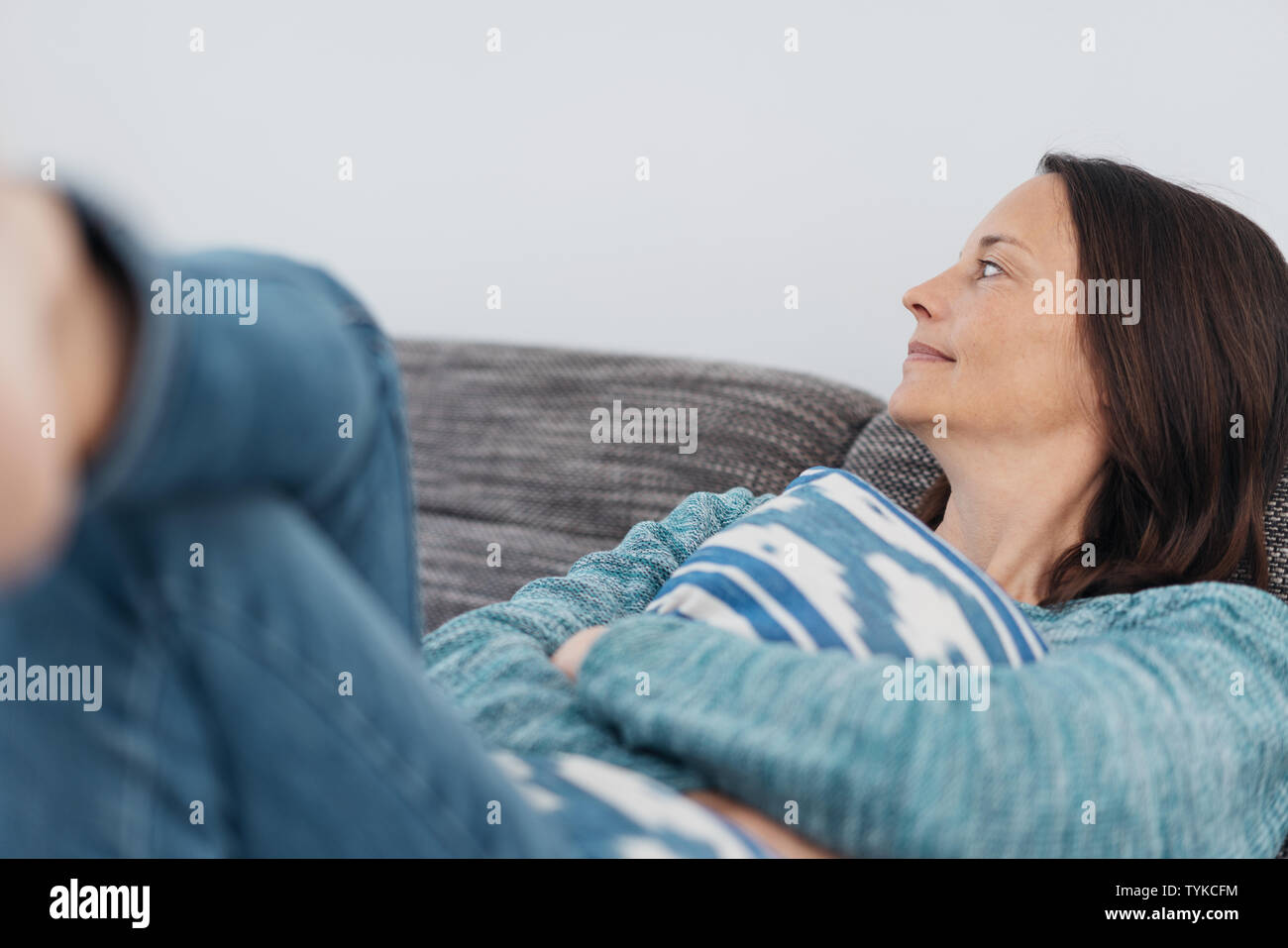 Adult smiling woman holding blue and white striped pillow with both arms while sitting back on couch Stock Photo