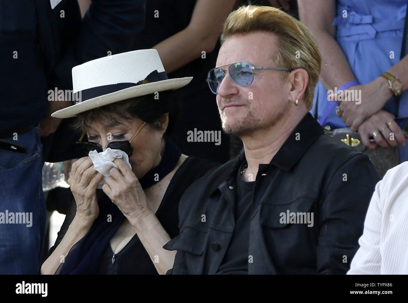 Bono of U2 sits with Yoko Ono who holds a tissue to her face as