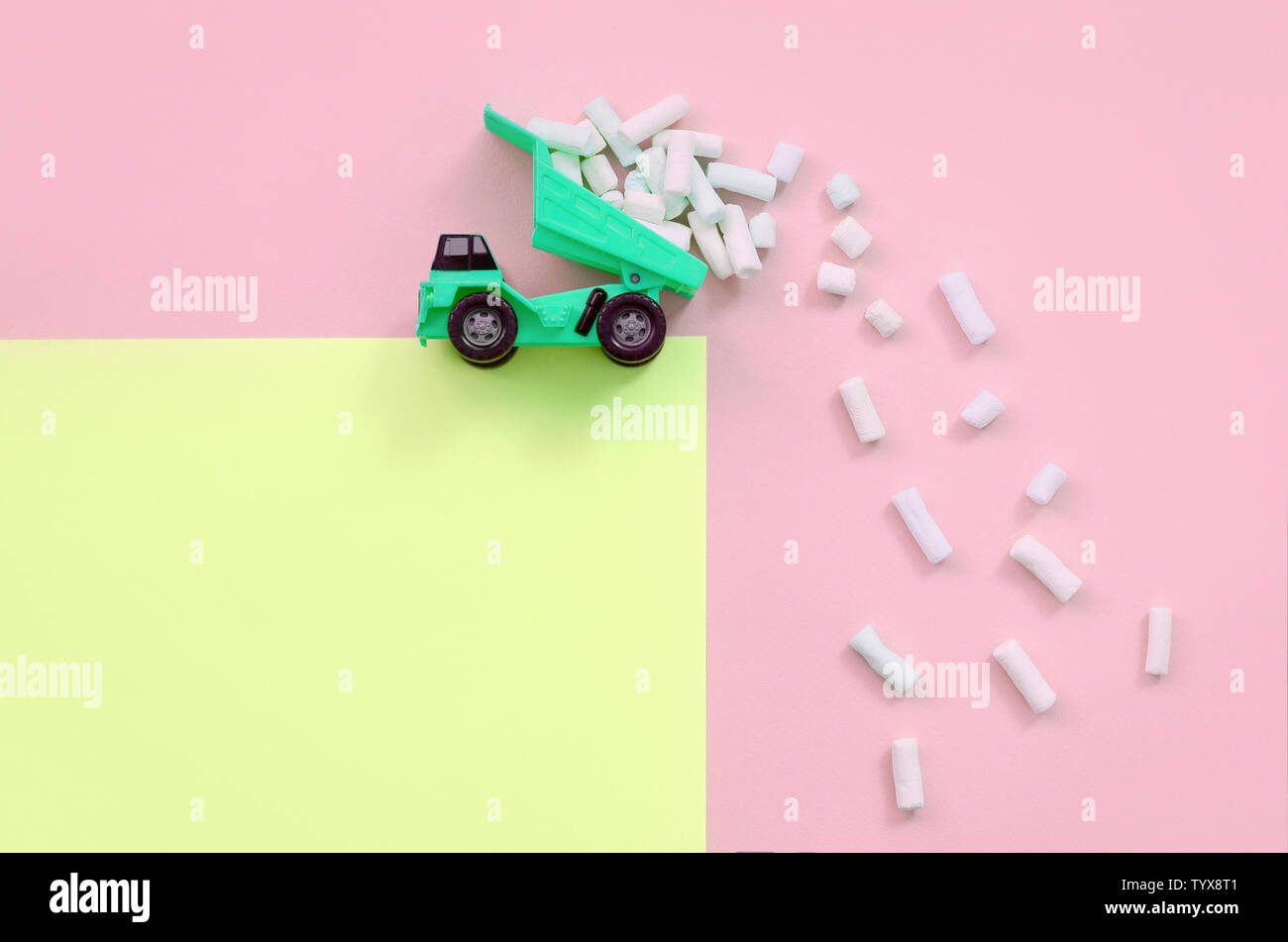 Green little toy dump truck throws marshmallow pieces from its raised back on a pastel yellow and coral background. Flat lay minimal top view. Stock Photo
