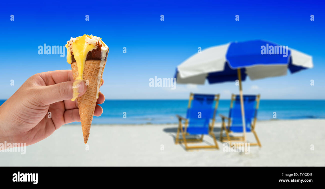 hand with a melting ice cream in front of blur sun loungers and a beach umbrella on silver sand Stock Photo