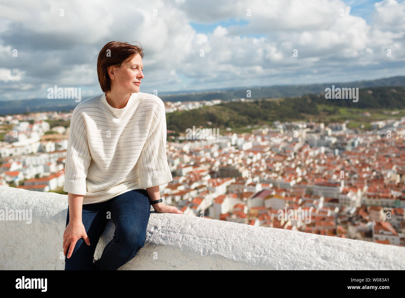 Adult woman sitting on stone fence and looking at cityscape of Nazare, Portugal Stock Photo