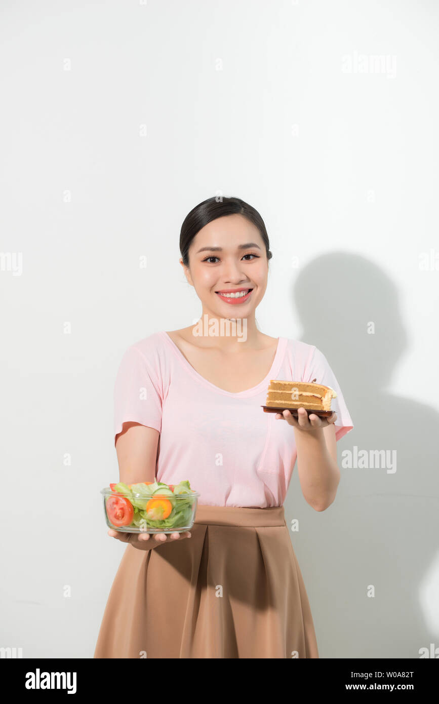 Half-length portrait of very beautiful woman holding small cake, fresh vegetables. Young housewife choosing sweets or healthy eating - cake and salad. Stock Photo