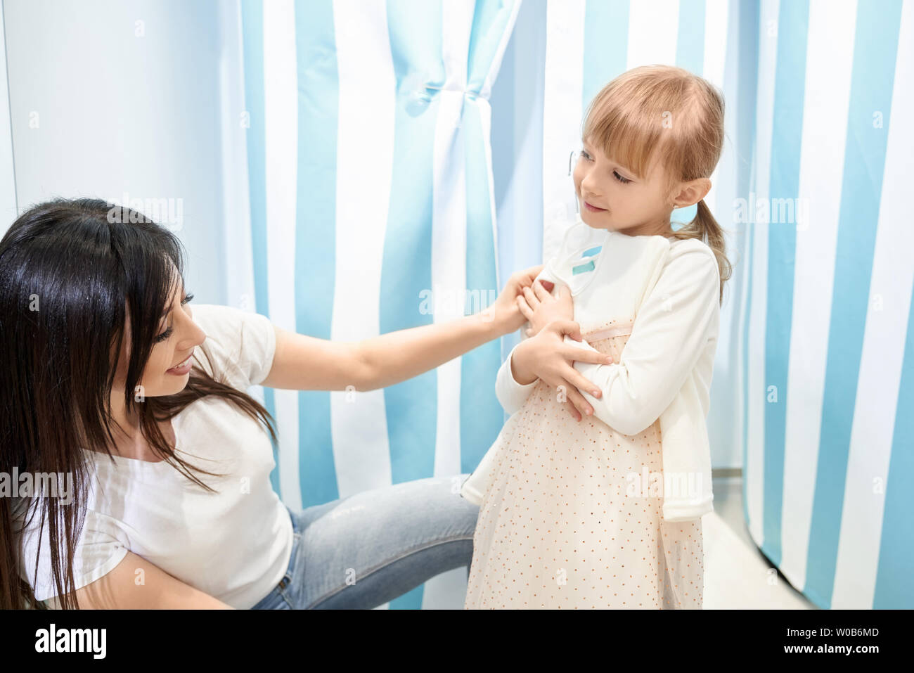 Smiling girl trying on new lovely dress in fitting room. Little daughter shopping together with beautiful mother choosing and buying outfit in stores. Concept of shopping time and dream Stock Photo