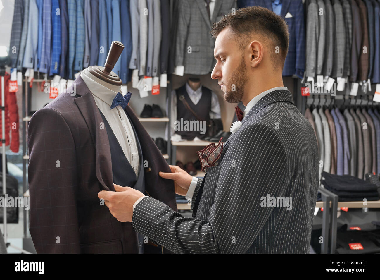 Young, handsome man choosing suit in fashionable boutique. Jackets of fashionable prints on hangers in row, clothing and footwear on shelves. Male clothing on mannequin. Stock Photo