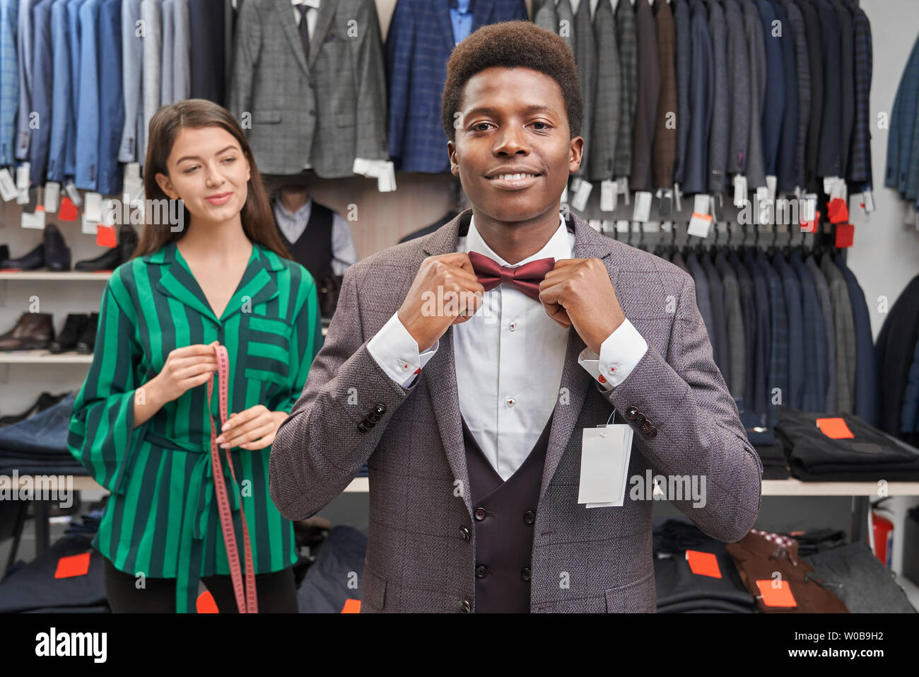 Client choosing suit, dressing white shirt, waistcoat and jacket, looking at camera and smiling. Handsome man with curly hair touching red bow tie, posing. Pretty seller holding measuring tape Stock Photo - Client choosing suit, dressing white shirt, waistcoat and jacket, looking at camera and smiling. Handsome man with curly hair touching red bow tie, posing. Pretty seller holding measuring tape Stock Photo -