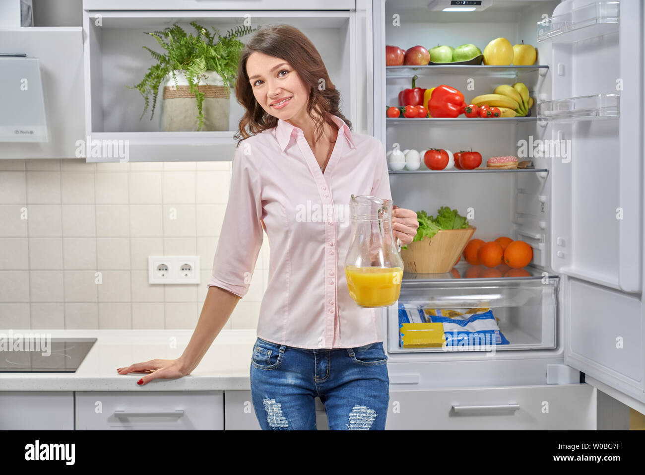 Front view of young attractive woman standing near opened refrigerator, keeping orange juice in hand, looking at camera and smiling. Healthy female drinking fresh juice at kitchen. Concept of health. Stock Photo