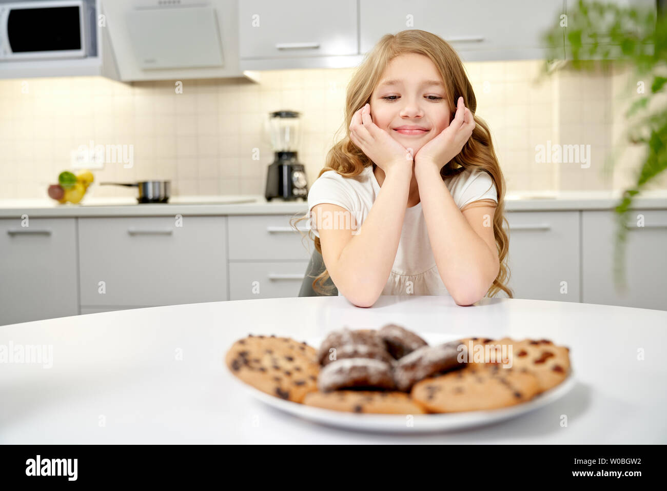 Front view of beautiful girl in white shirt sitting at table in kitchen, putting head on hands and looking at tasty cookies. Pretty child wishing chocolate desserts. Concept of sweets and eating. Stock Photo