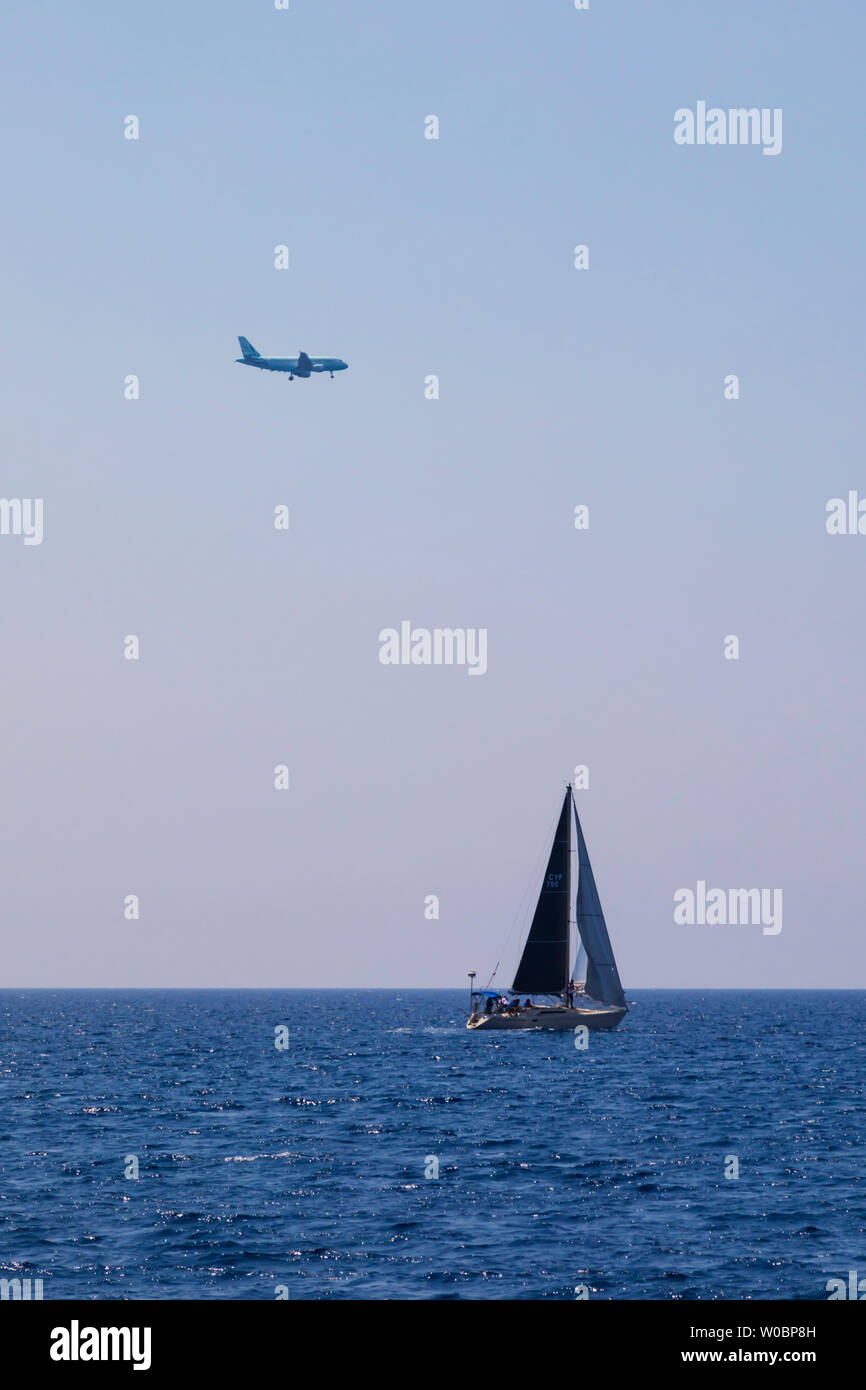 An airliner on approach to Larnaca airport passes over a yacht at sea. Larnaca,  Cyprus. June 2019 Stock Photo