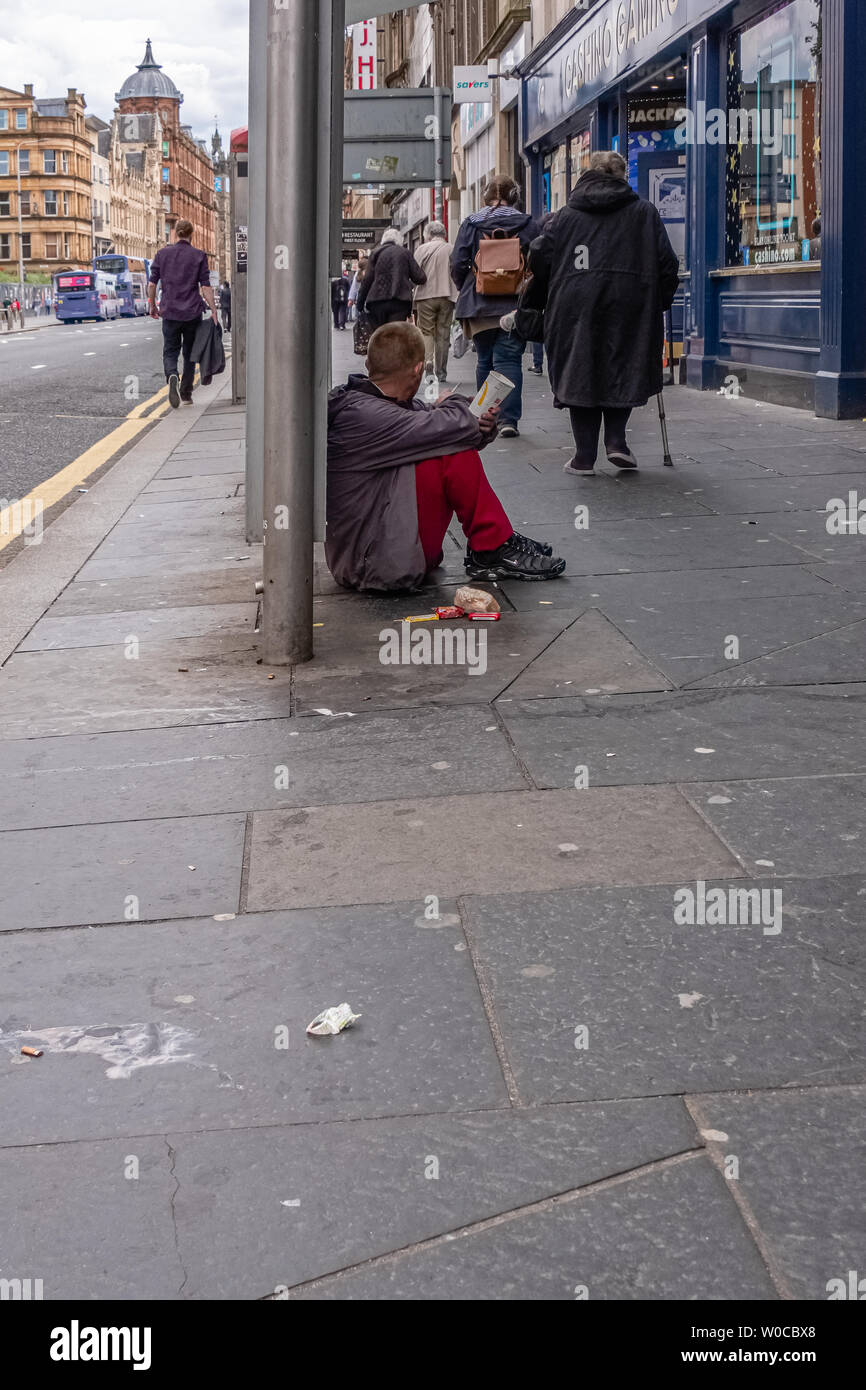 Glasgow, Scotland, UK - June 22, 2019: People walking past a male gentleman siting on the dirty pavement with a McDonald's paper cup appealing for mon Stock Photo