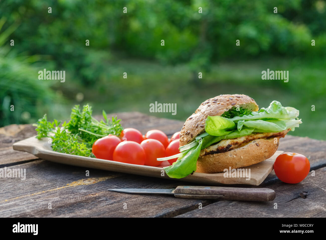 Tasty homemade burger with meat, lettuce, tomatoes, sesame bun on picnic table in garden outdoors. Stock Photo