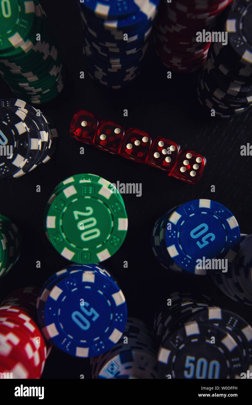 Close up stacks of different colored poker chips and playing dices on the casino table isolated over black background. Gambling tournament betting for Stock Photo