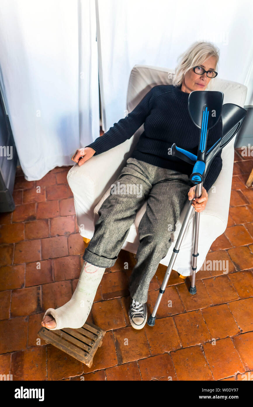 Woman sat in a chair with one plastered-leg on a footrest and holding her crutches Stock Photo