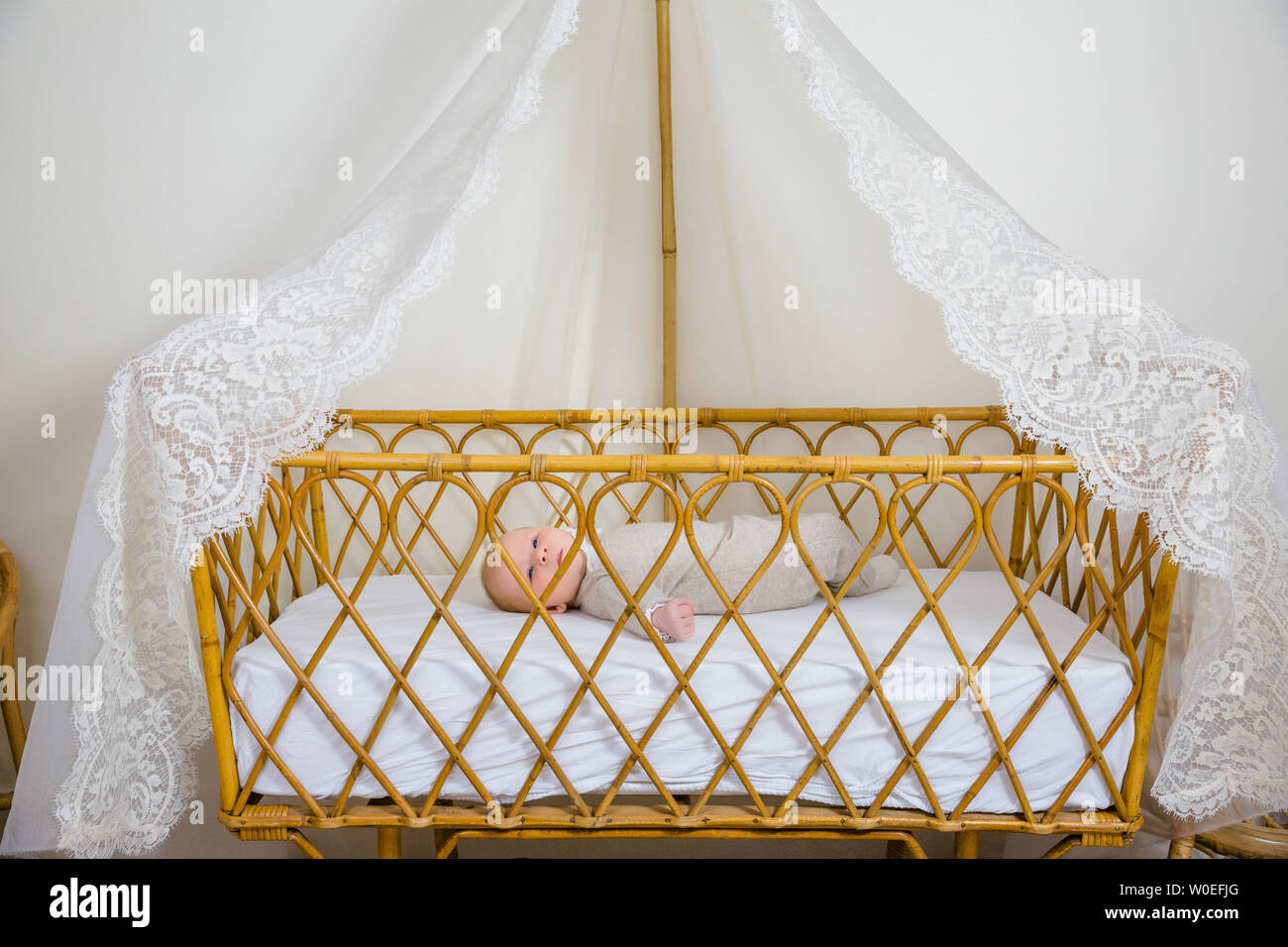 View of a baby awake 2 months in beige layette lying on his back in his baby bed canopy wicker and lace. Stock Photo