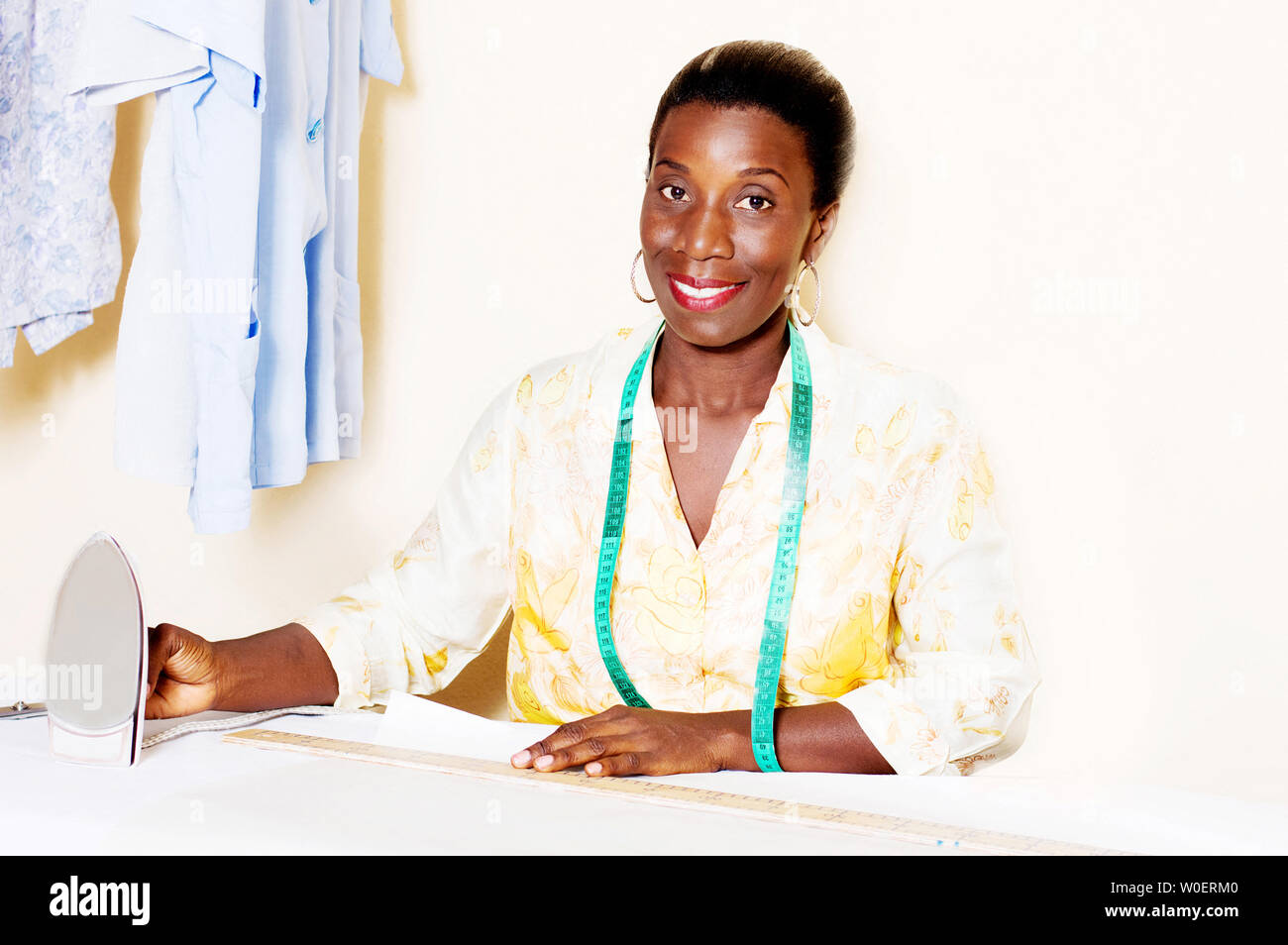 Young smiling seamstress holding an iron in front of her work table. Stock Photo