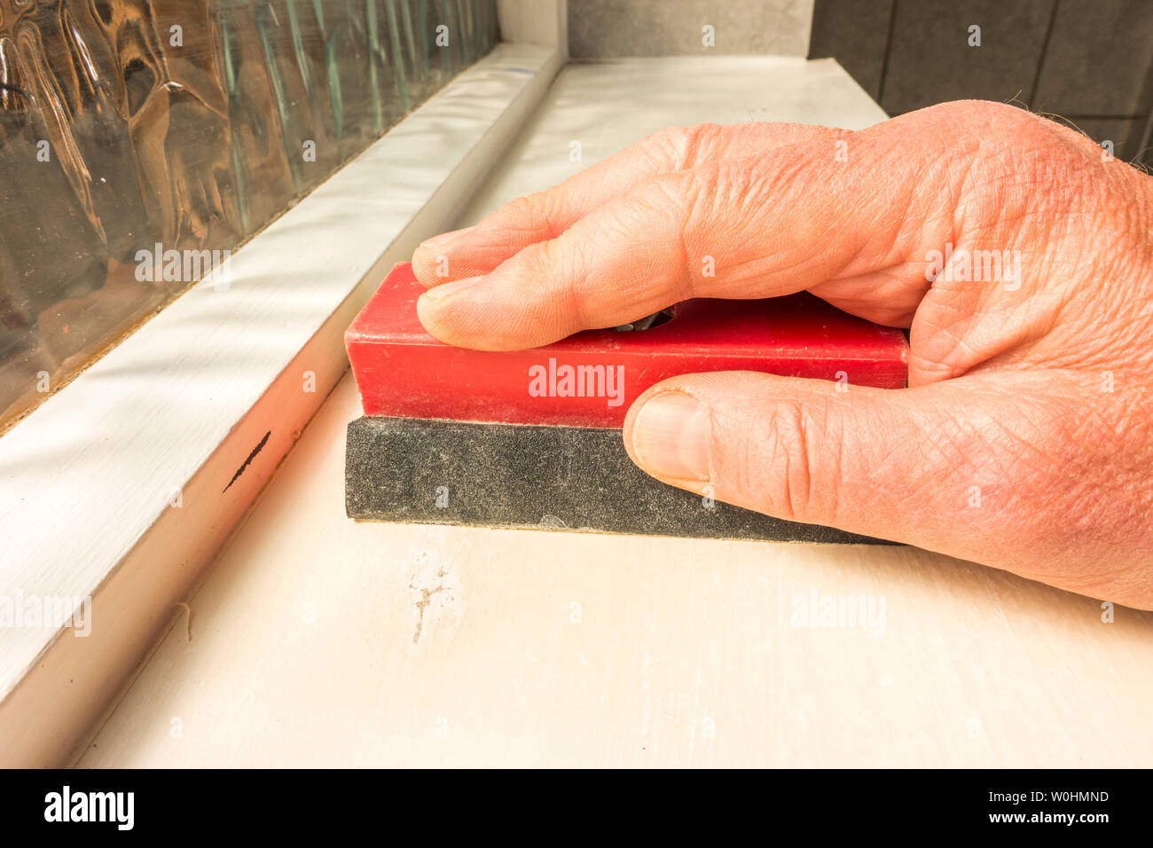 Man’s hand using a sanding block with abrasive sandpaper, to rub down a painted bathroom shelf prior to repainting. Stock Photo