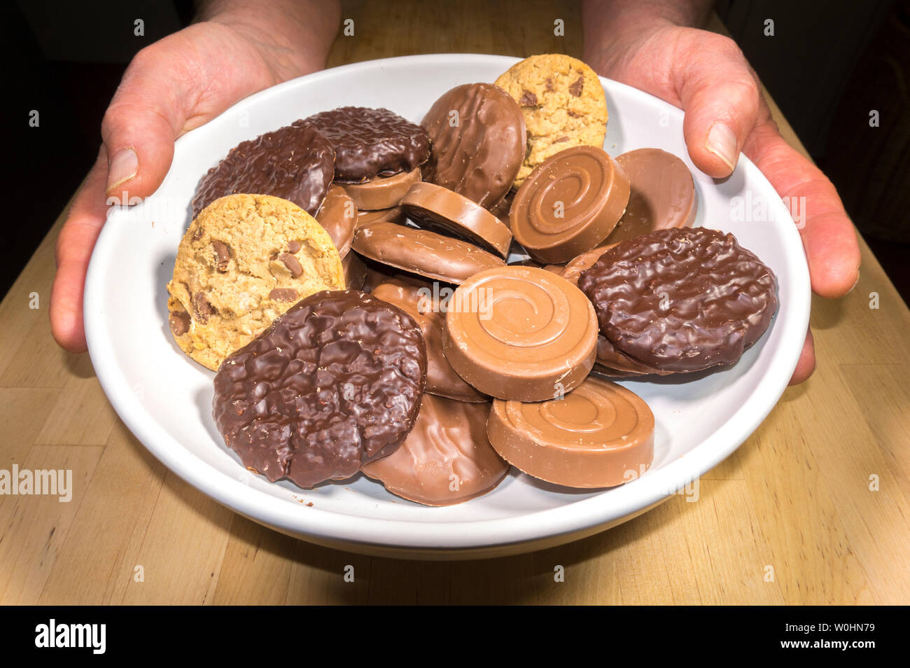 Closeup of a man’s hands serving a white shallow bowl on a table, containing a variety of round chocolate coated biscuits. Stock Photo