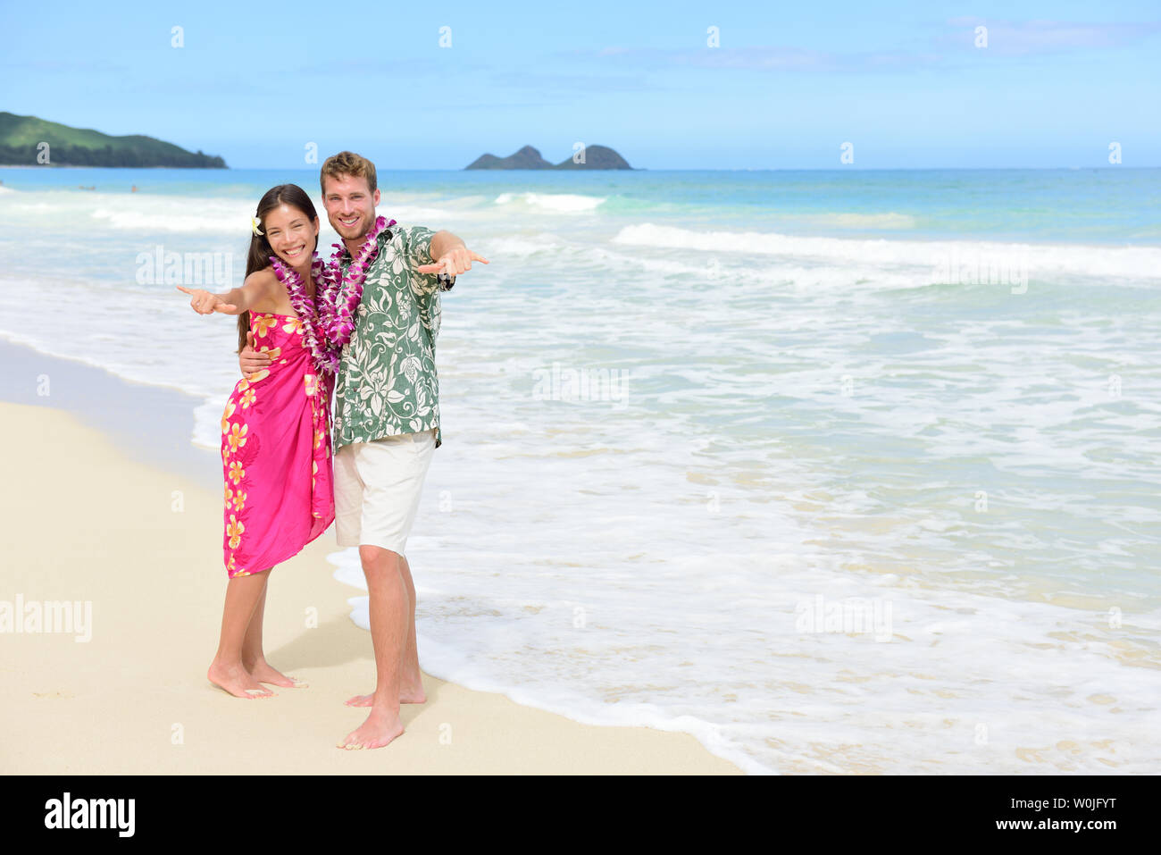 Portrait of couple of tourists happy standing on Hawaiian beach at their Hawaii vacation. Asian woman and Caucasian man wearing flower lei garland and Aloha clothing showing Shaka hand sign on travel. Stock Photo