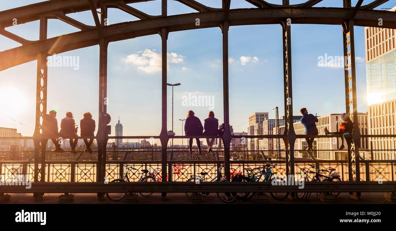 Young people sitting on the balustrade of the Hackerbrucke bridge over the railway tracks and looking into the distance, back light, Munich, Upper Stock Photo