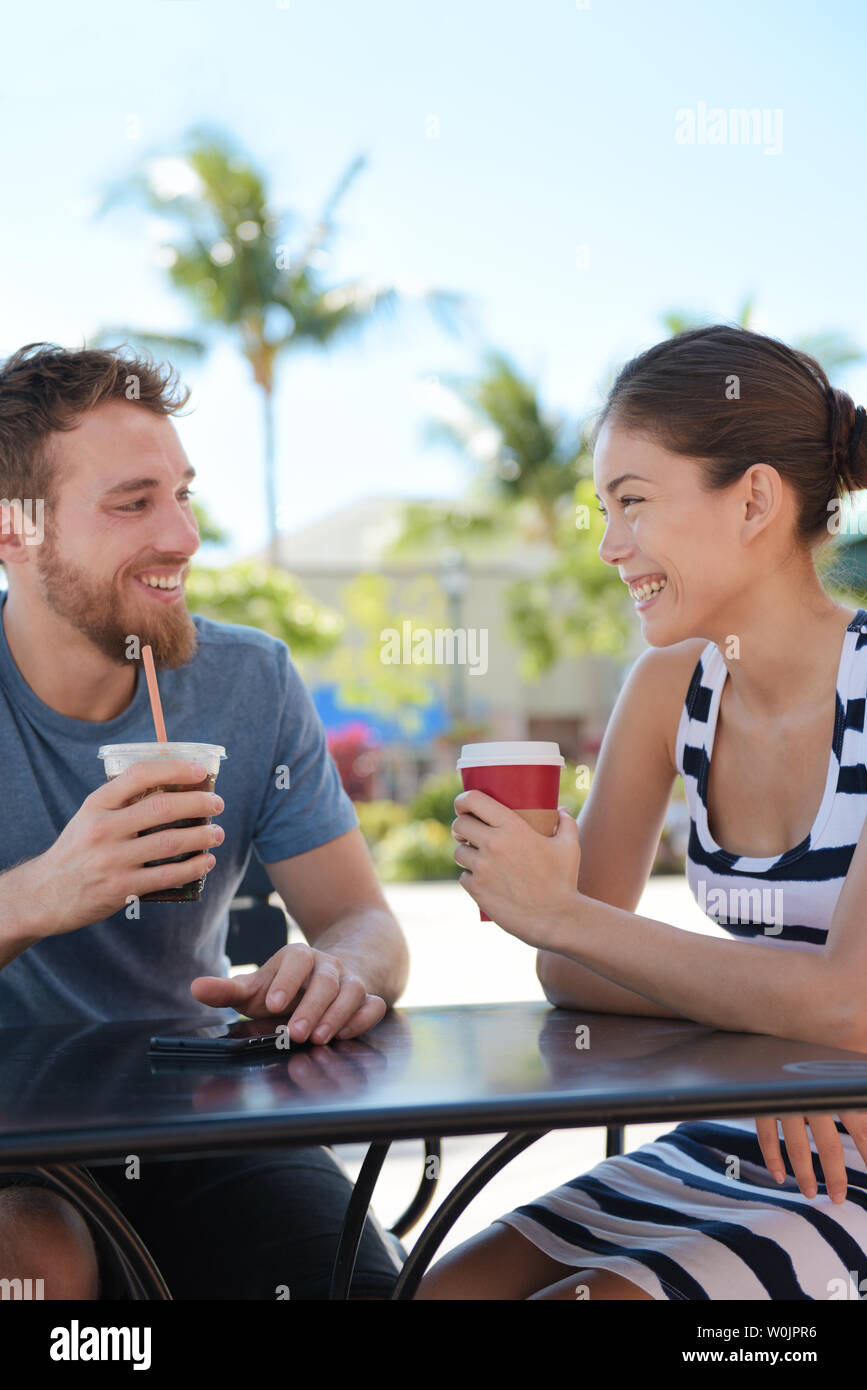 Cafe couple having fun drinking coffee talking smiling and laughing on date in summer. Young man talking with Asian woman sitting outdoors. Happy friends in late 20s. Stock Photo