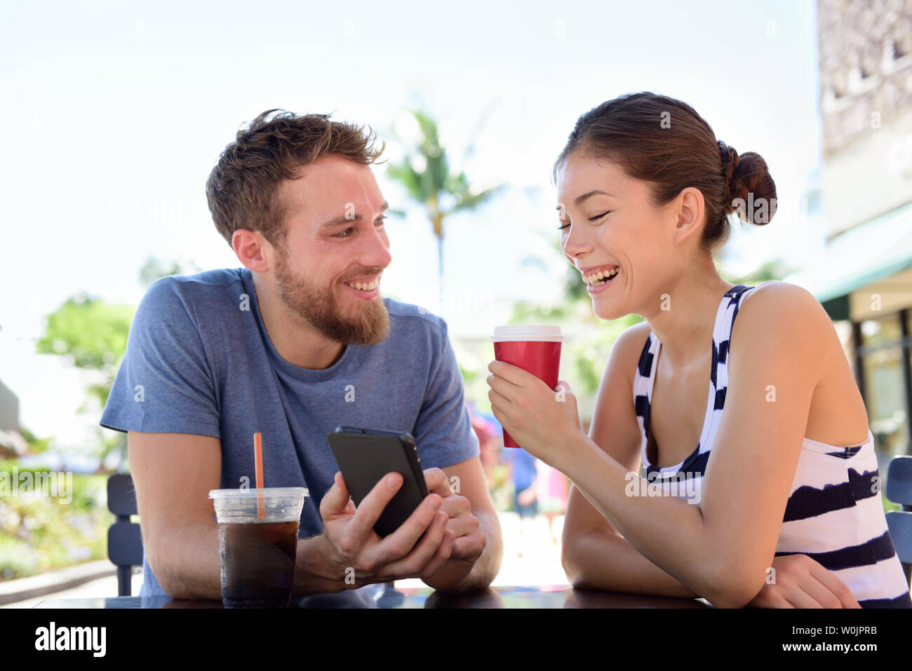 Couple on cafe looking at smart phone app pictures drinking coffee in summer. Young urban man using smartphone smiling happy to casual asian woman sitting outdoors. Friends in late 20s Stock Photo