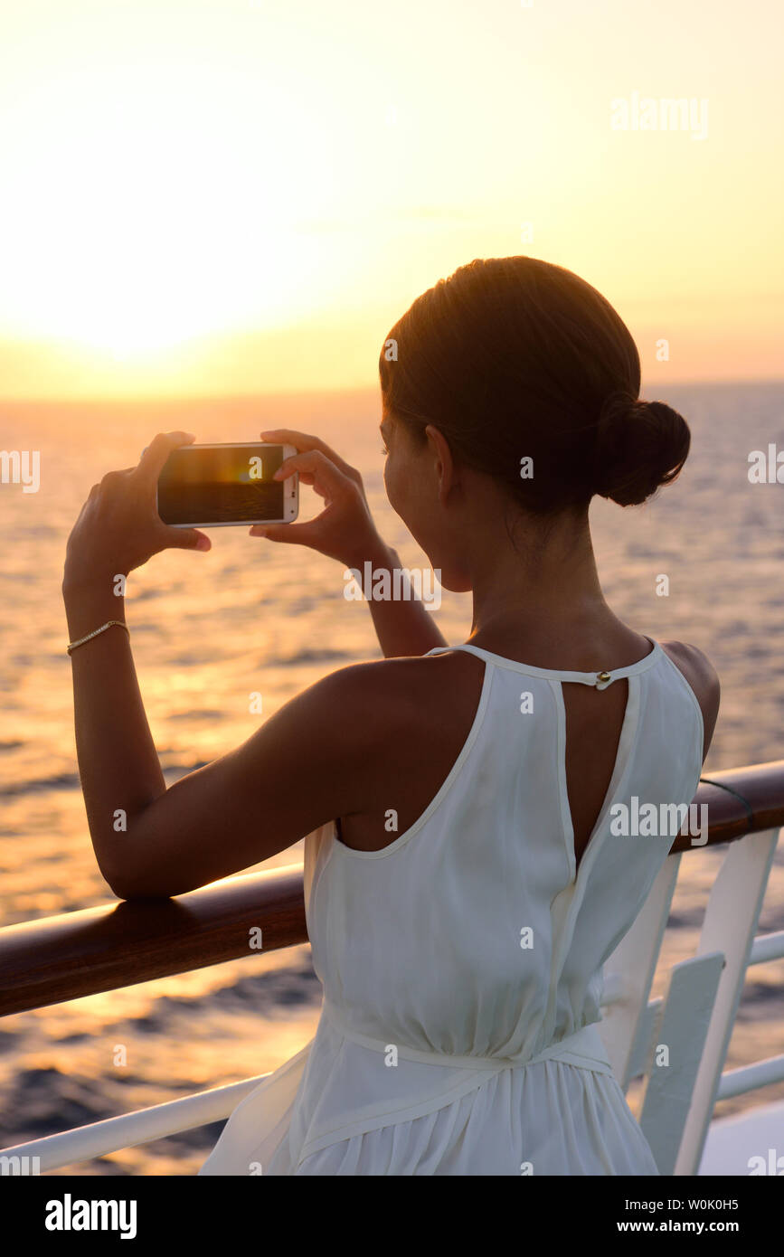 Cruise ship vacation woman taking photo with smart phone camera enjoying sunset on travel at sea. Girl using smartphone to take picture of ocean sunset. Woman in dress on luxury cruise liner boat. Stock Photo