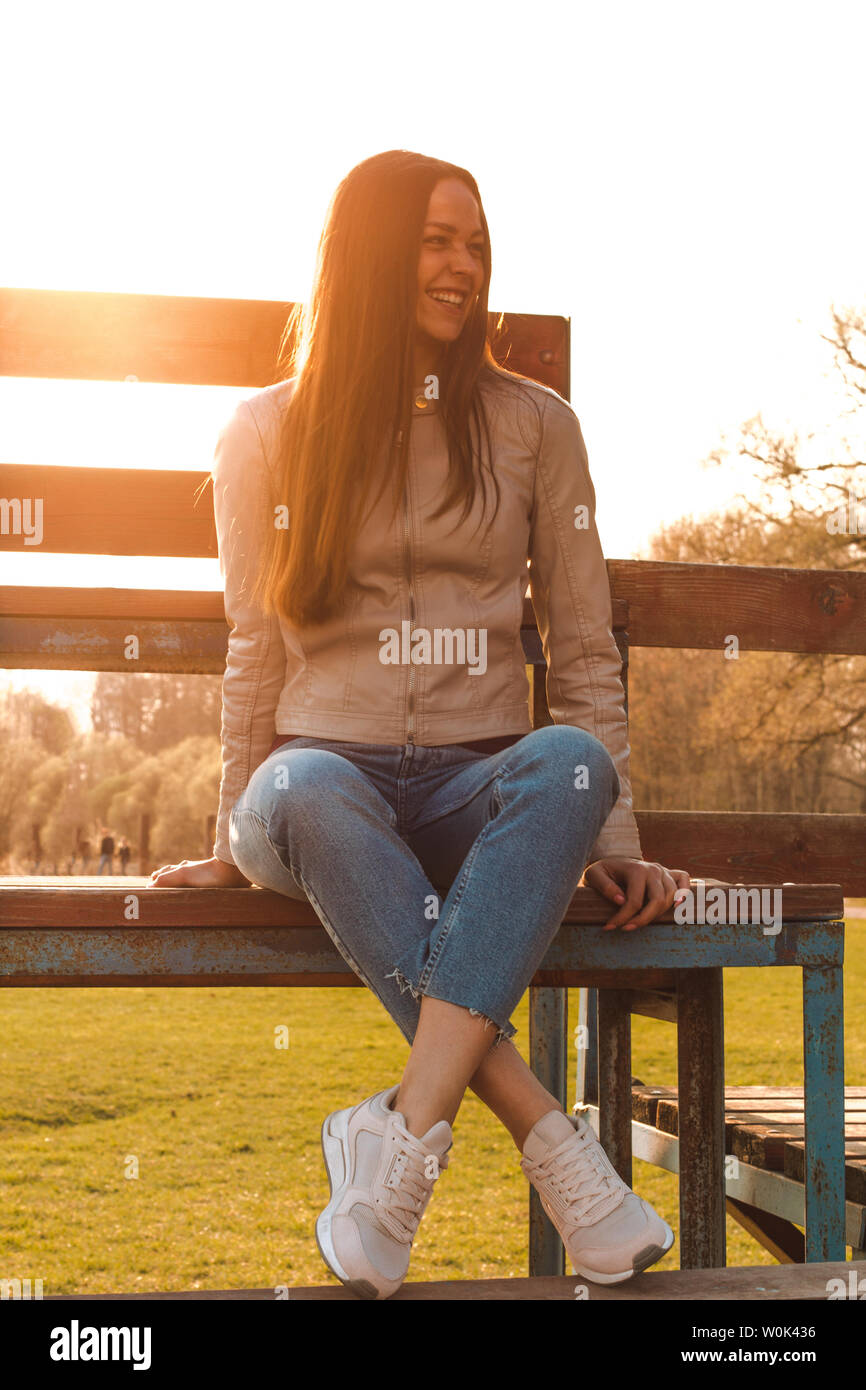 beautiful girl with long hair sits opposite the sunlight in a beige leather  jacket, jeans and sneakers laughing a lot Stock Photo - Alamy
