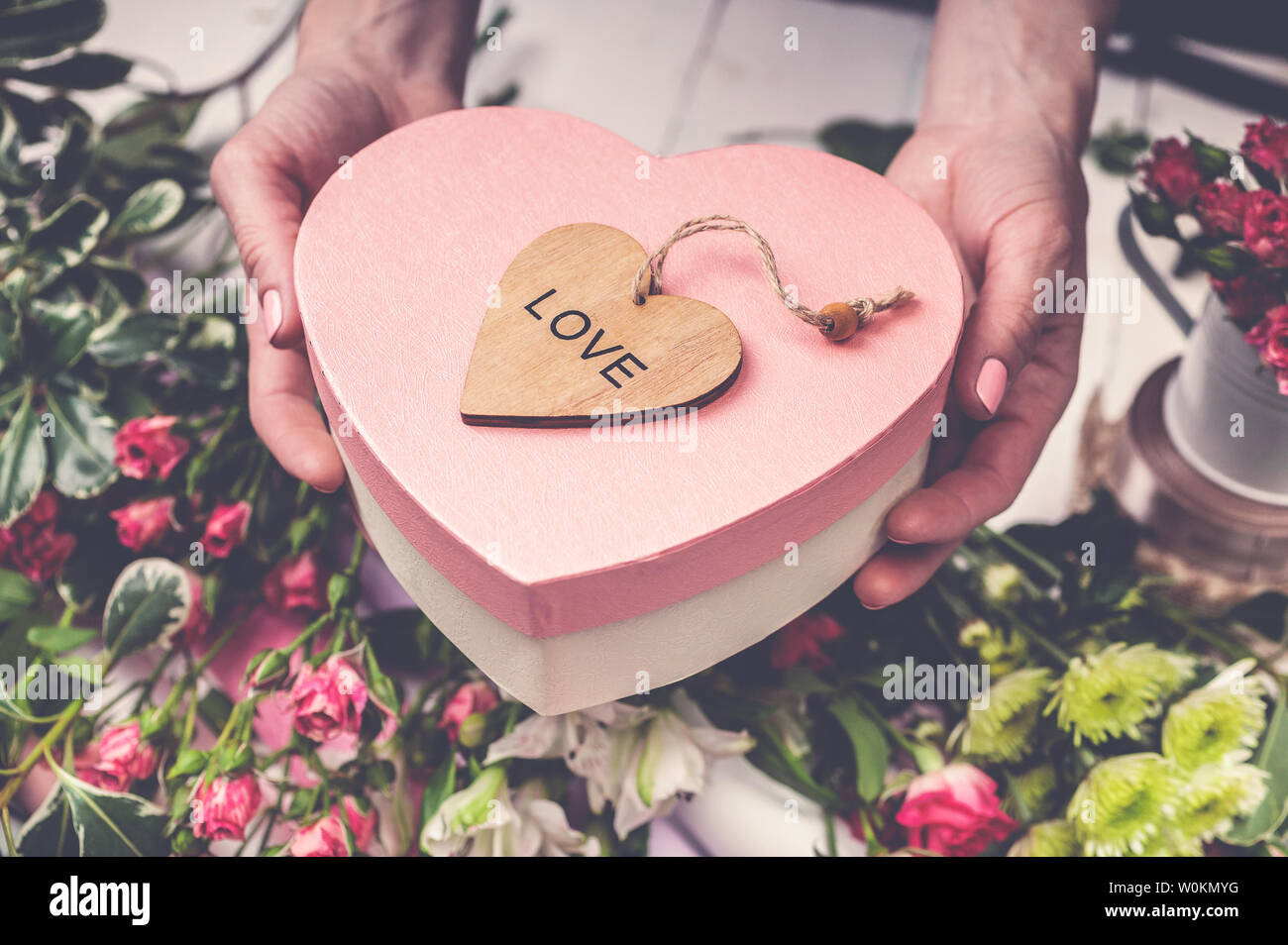 Florist's workplace: flowers, accessories, tools. Female hands holding a gift box in the shape of a heart. Stock Photo