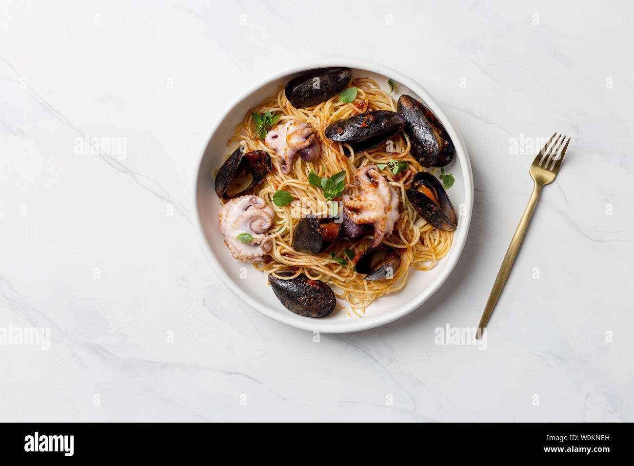 Italian pasta with mussels and octopus in white plate with golden fork on white marble background. Concept of Italian dinner Stock Photo
