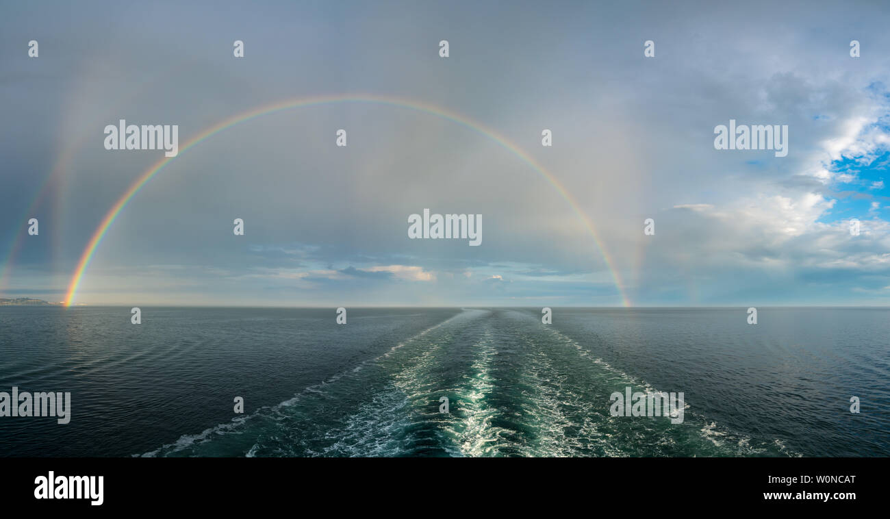 Dramatic double rainbow forms over the wake of a cruise ship at sea Stock Photo